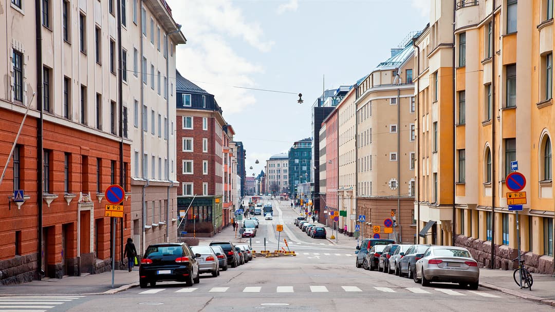 katukuva helsingistä. kadunvarrella näkyy eri aikakausina rakennettuja betonisia kerrostaloja. Tienviereen on parkkeerattu autoja